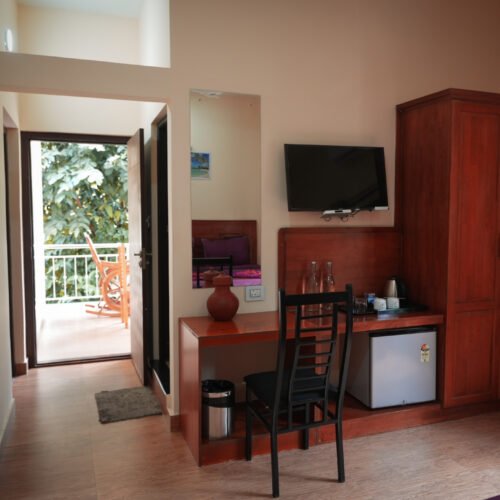 A cozy hotel room with wooden furniture, including a desk with a chair, a wall-mounted TV, a wardrobe, and a mini fridge. The room has a balcony view with a wooden rocking chair surrounded by lush greenery.